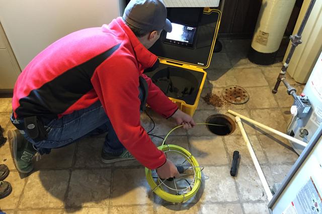 Plumber in a red jacket feeding a drain inspection camera cable into a floor drain